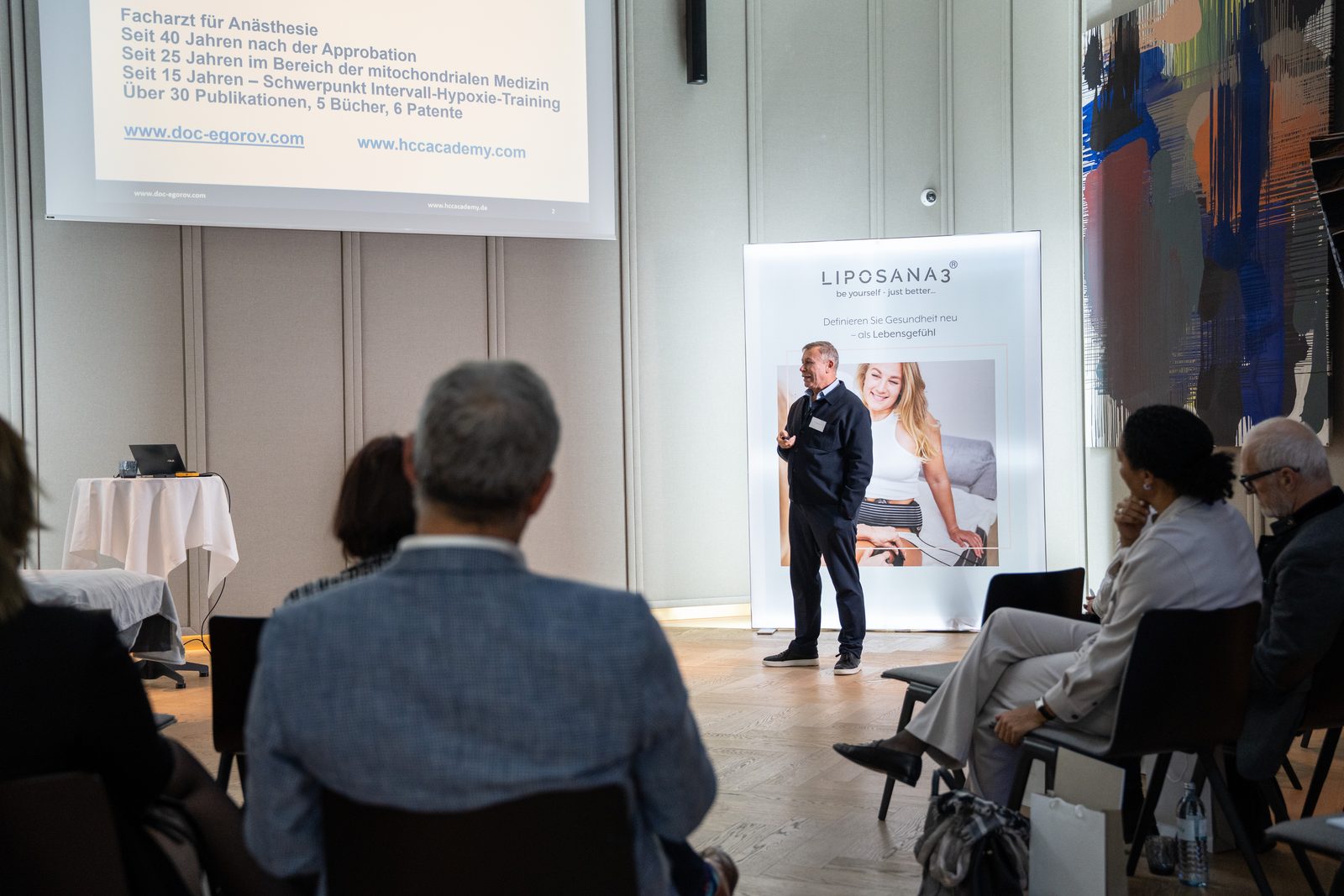 Ein Arzt spricht vor Publikum, auf der Leinwand seine Qualifikationen: Facharzt seit 40 Jahren, über 30 Publikationen, 5 Bücher, 6 Patente